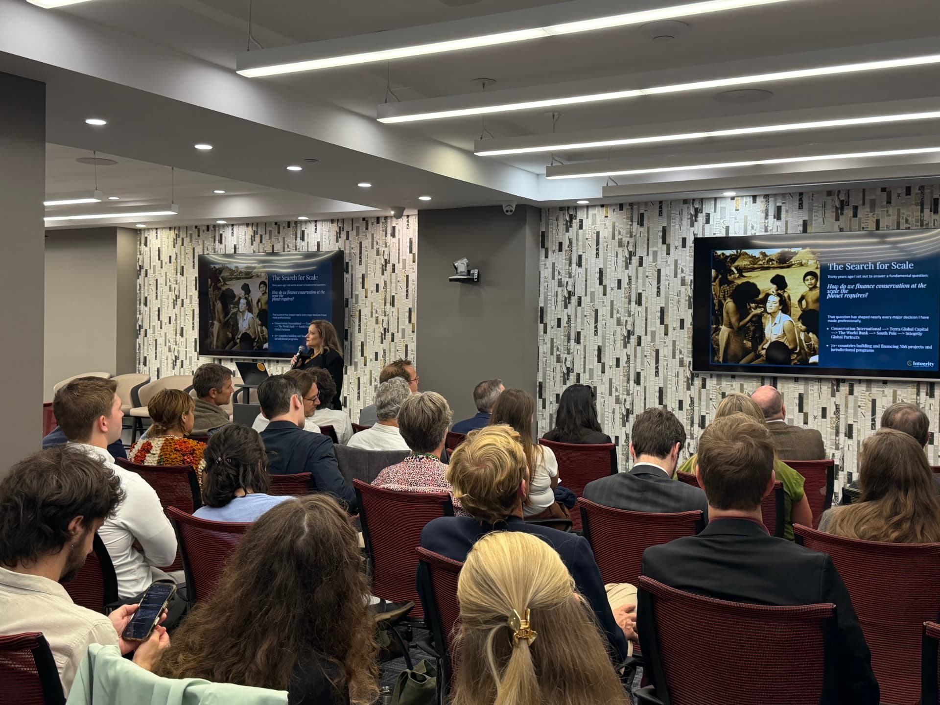 A speaker addressing a seated audience at an Integrity-hosted session, with the slide 'The Search for Scale' shown on two screens